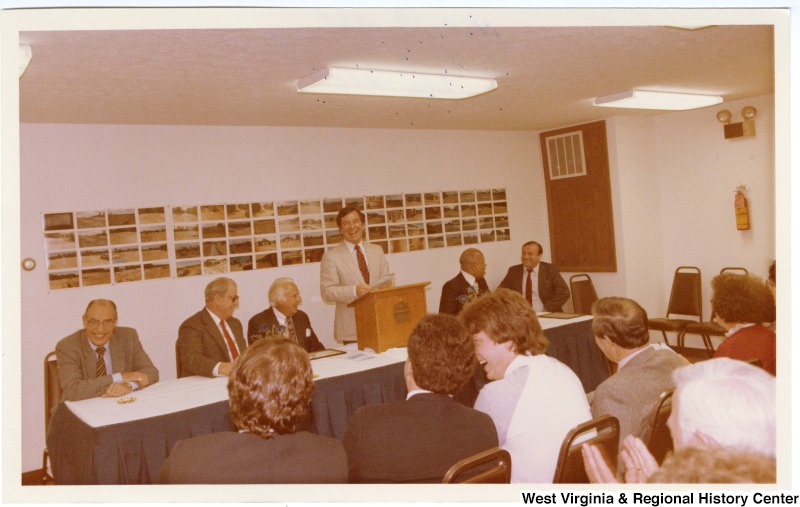 Congressman Nick Rahall, II speaking at a Beckwood Housing Project meeting in Beckley, West Virginia. Five unidentified men are on the stage with him.