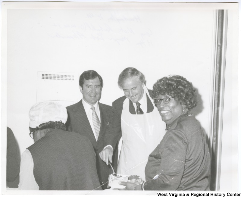 Congressman Nick Rahall, II (left) and Huntington Mayor Robert "Bob" P. Alexander serving food at a community event. Two unknown women are in the foreground of the photo.