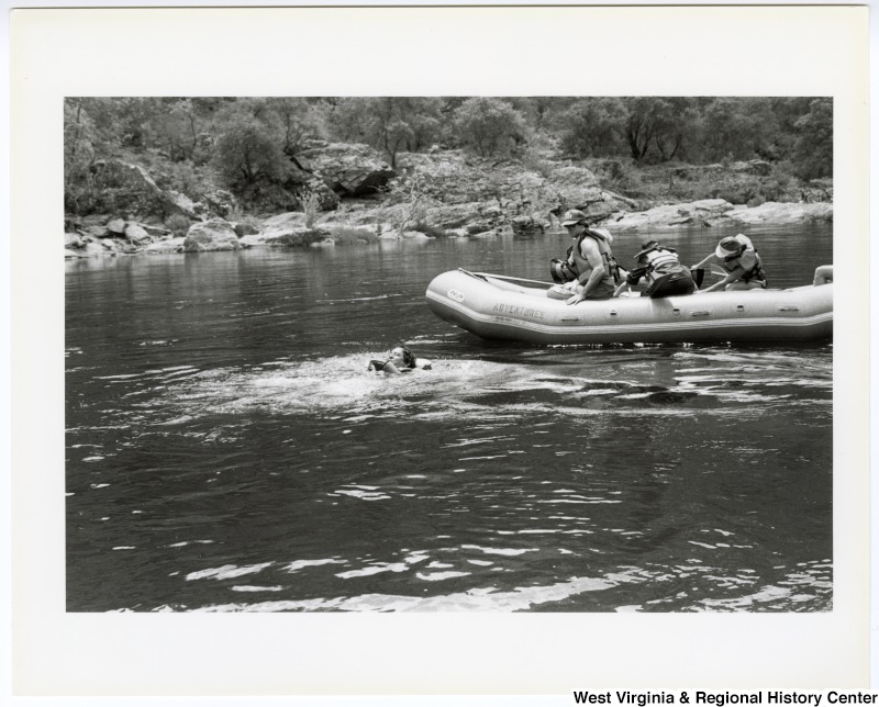 Congressman Nick Rahall (left) swimming in the Tuolumne River in central California during a whitewater rafting trip. Three unidentified people sit in the raft to the right.