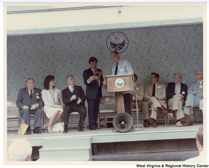 Congressman Nick Rahall, II being handed a plaque at the Beckley sewage treatment plant groundbreaking. Senators Jennings Randolph (left) and Robert C. Byrd (third from left) sit on stage.