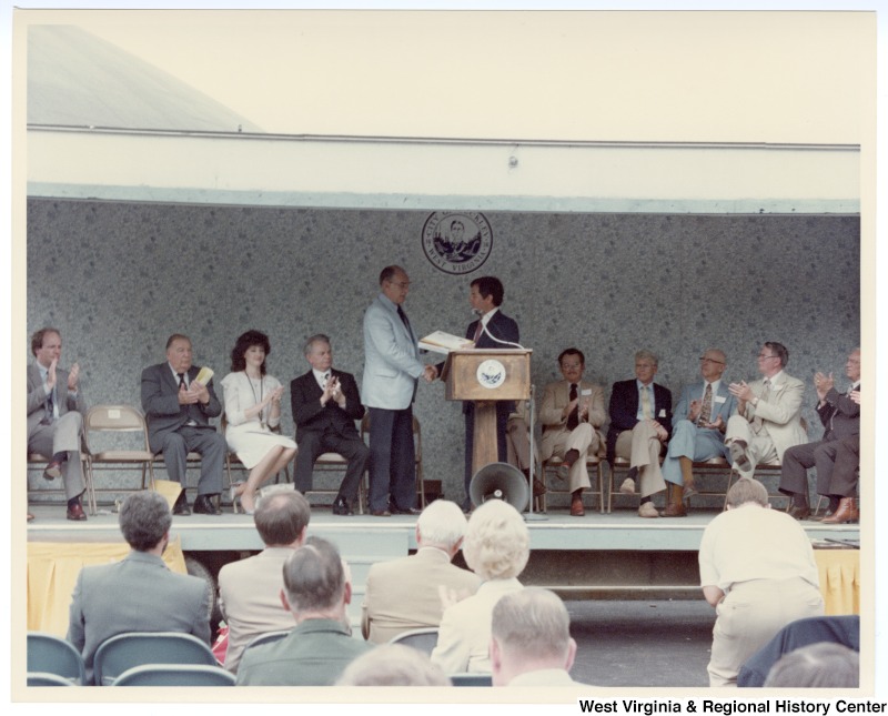 Congressman Nick Rahall, II at the podium with an unidentified man at the Beckley sewage treatment plant groundbreaking. Senators Jennings Randolph (second from left) and Robert C. Byrd (fourth from left) sit on stage.