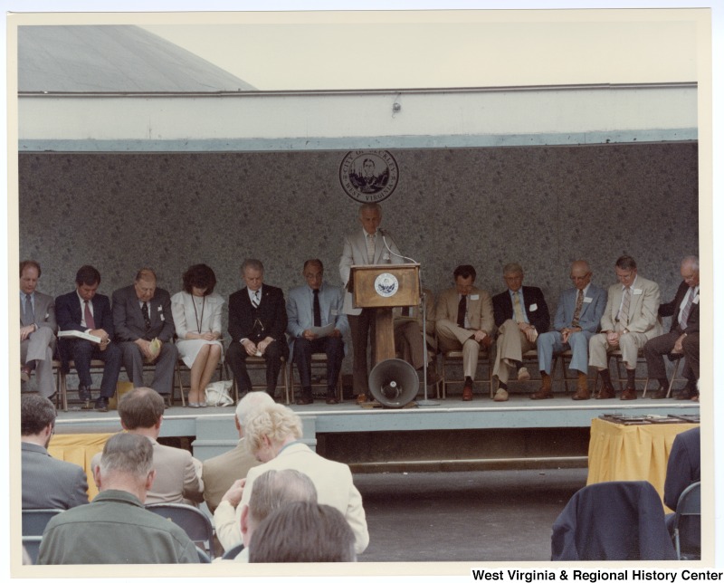 An unidentified man speaking at the groundbreaking for the Beckley sewage treatment plant. Seated behind the unidentified man is Congressman Nick Rahall, II (second from left) and Senators Jennings Randolph (third from left) and Robert C. Byrd (fifth from left).