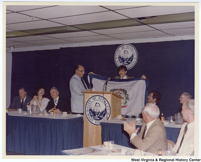 Senators Jennings Randolph (left in back row) and Robert C. Byrd (third from left in back row) as well as Congressman Nick Rahall, II (second from right in back row) at a banquet following the groundbreaking of the Beckley Sewage Treatment plant. An unidentified man holds the West Virginia flag.