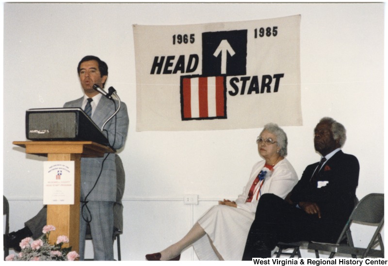 Congressman Nick Rahall, II speaking at McDowell County Head Start Program. Behind him on the stage sit an unidentified woman and man.The back of the image has written on it: "Lester Toney Bx III Amonate Va. 24601"