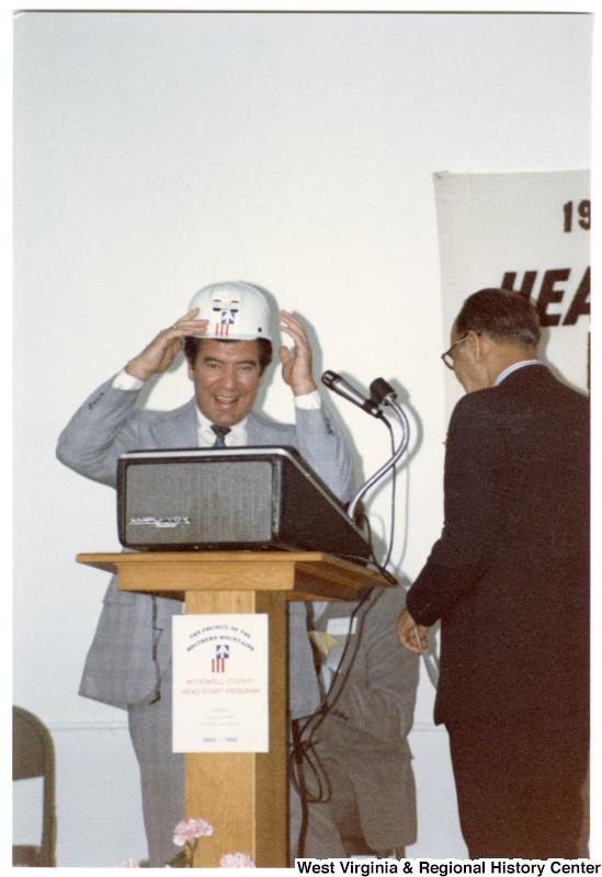 Congressman Nick Rahall, II speaking at McDowell County Head Start Program. He is placing a Head Start hard hat on his head. Behind him on the stage sits an unidentified man and to Rahall's right is another unidentified man.The back of the image has written on it: "Lester Toney Bx III Amonate Va. 24601"