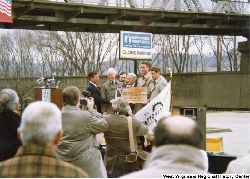 Congressman Nick Rahall II (left), Senator Robert C. Byrd (second from left), with four unidentified men holding a check for $20,000,000 that will replace or repair the Huntington Sixth Street Bridge. The men stand in front of the bridge.