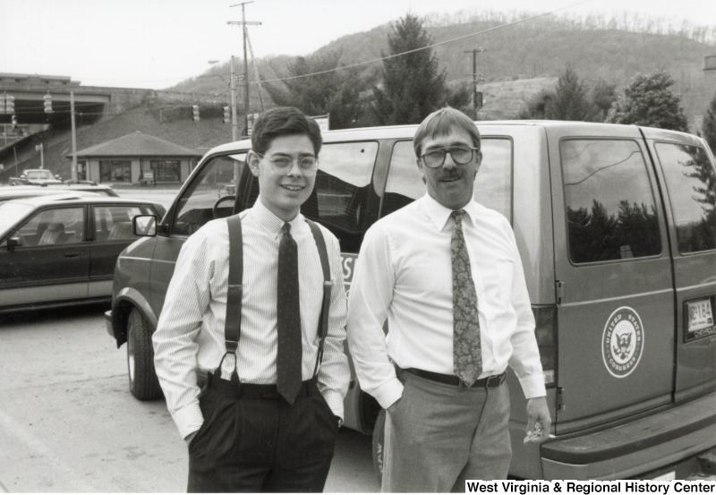 Two unidentified men standing in front of a van with a United States Congress logo affixed to it.