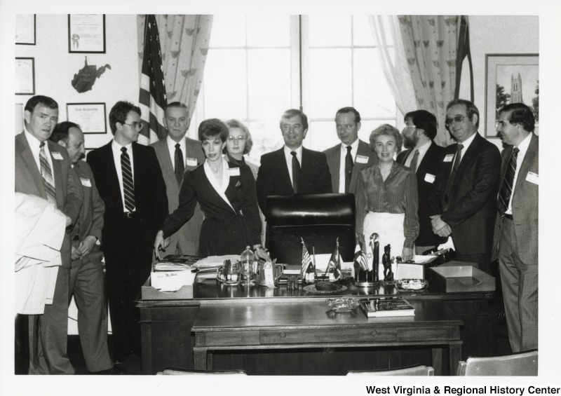 Representative Nick J. Rahall standing behind the desk in his office surrounded by a large group of unidentified men and women