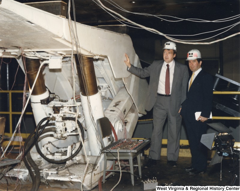 Representative Nick J. Rahall (D-W.Va.) with an unidentified man standing beside mining machinery equipment.