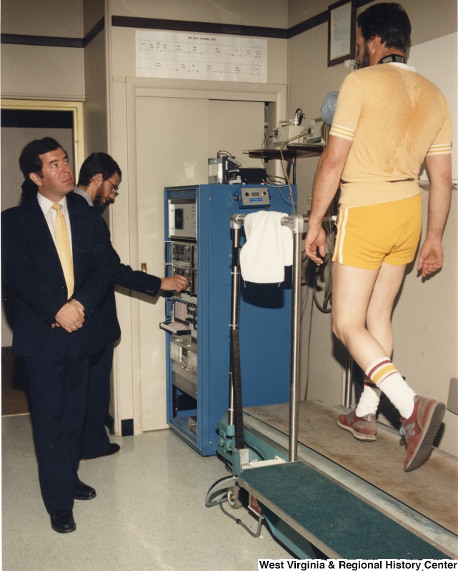 Representative Nick J. Rahall (D-W.Va.) stands next to an unidentified man on a treadmill. An unidentified man stands behind Rahall adjusting a machine.