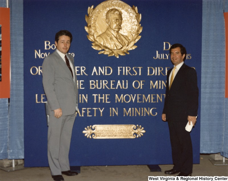 Representative Nick J. Rahall (D-W.Va.) stands next to an unidentified man in front of a commemorative wall honoring Joseph Austin Holmes and mining safety.