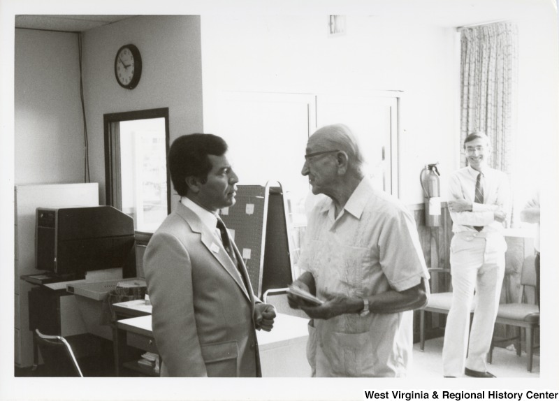 On the left, Representative Nick J. Rahall (D-W.Va.) speaks to an unidentified older man. In the background an unidentified man is standing watching them talk.