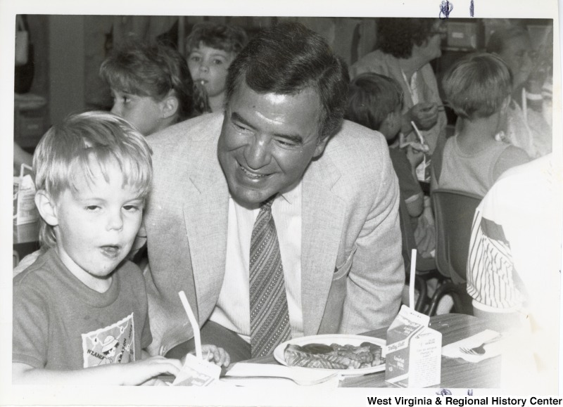 Representative Nick J. Rahall (D-W.Va.) seated next to an unidentified young boy eating a meal.