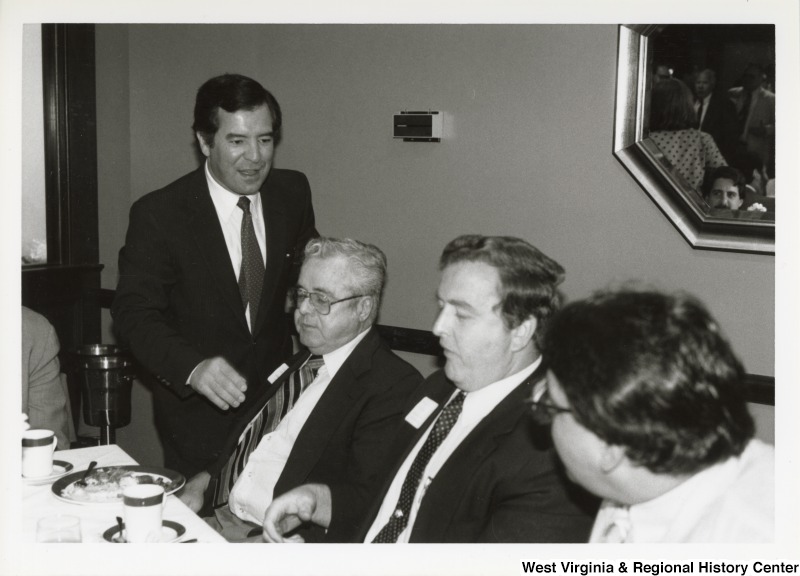 On the left, Representative Nick J. Rahall (D-W.Va.) talks with three unidentified, seated men at an event.