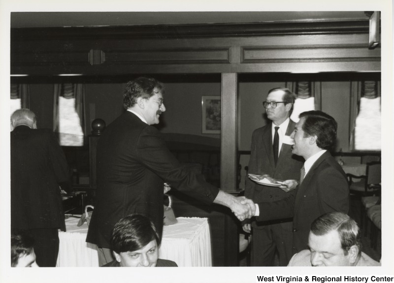 On the right, Representative Nick J. Rahall (D-W.Va.) shakes hands with an unidentified man at an event.
