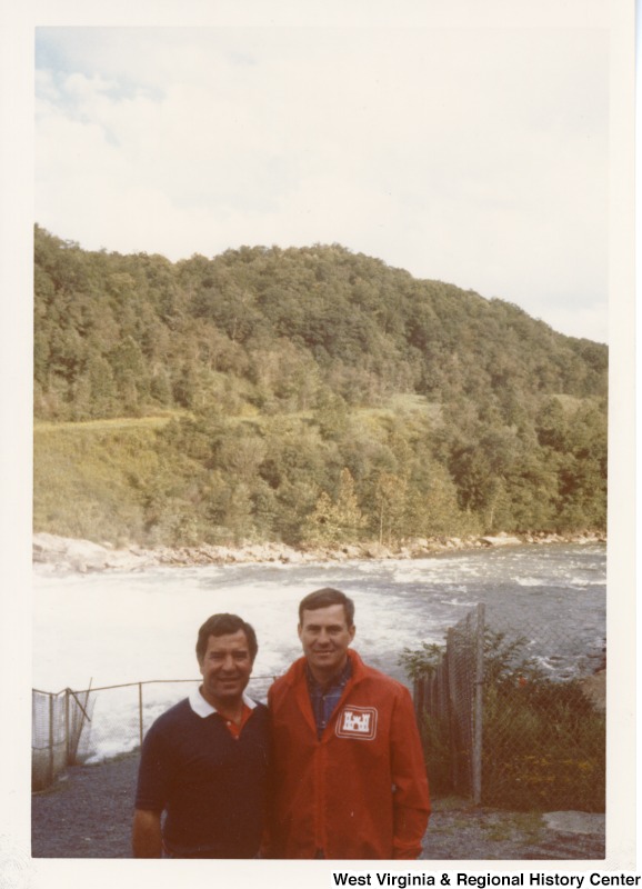 Representative Nick J. Rahall (D-W.Va.) stands beside an unidentified man at the edge of a river.