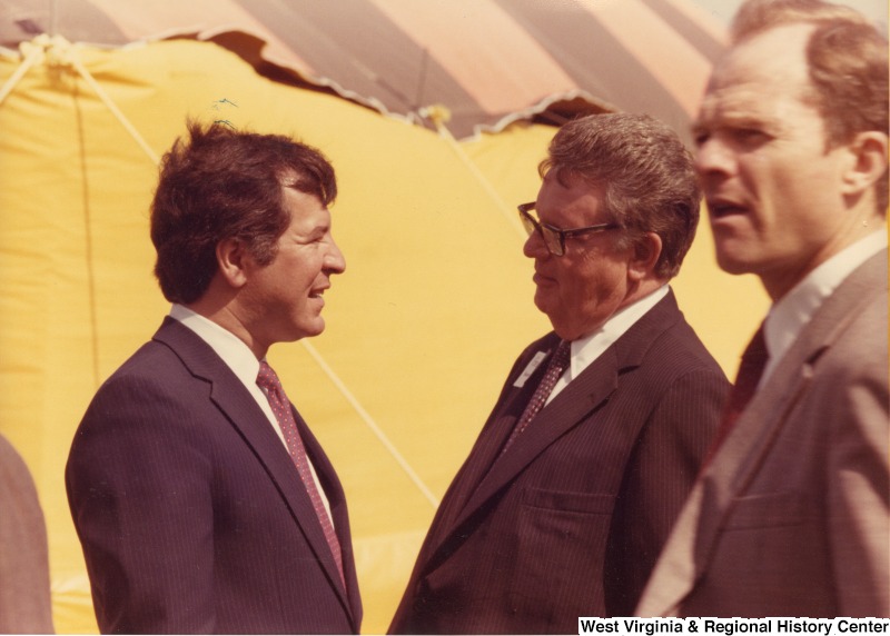 On the left, Representative Nick J. Rahall (D-W.Va.) talks with two unidentified men at an event outdoors.