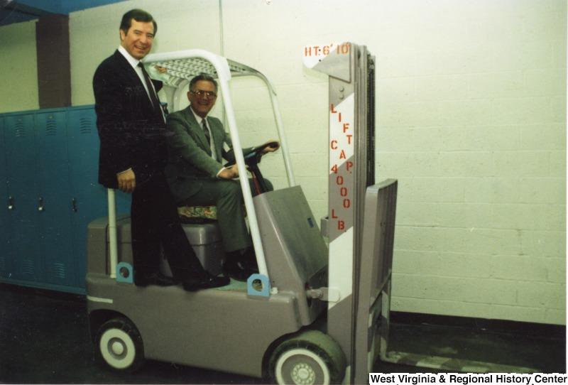 Representative Nick J. Rahall (D-W.Va.) stands on the side of a forklift operated by an unidentified man while getting a tour of Shawnee Hills Mental Retardation Center.