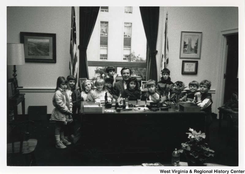 Seated at his desk, Representative Nick J. Rahall (D-W.Va.) is surrounded by an unidentified group of young children in an office room.