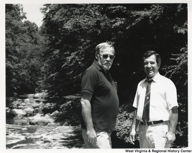 On the right, Representative Nick J. Rahall (D-W.Va.) smiles for a photograph with an unidentified man in front of a creek.