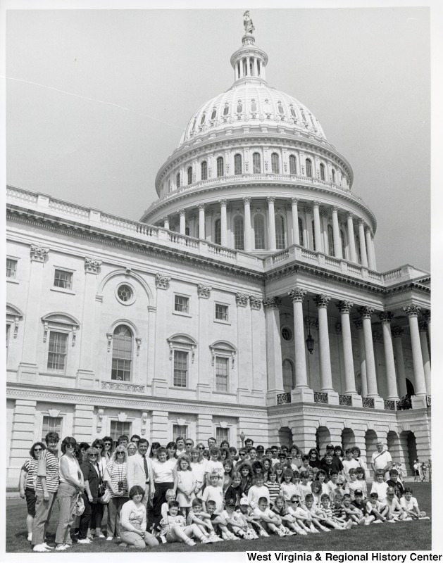 A large group of school children stands for a photograph in front of the United States Capitol building. Towards the left of the group stands Representative Nick J. Rahall (D-W.Va.).