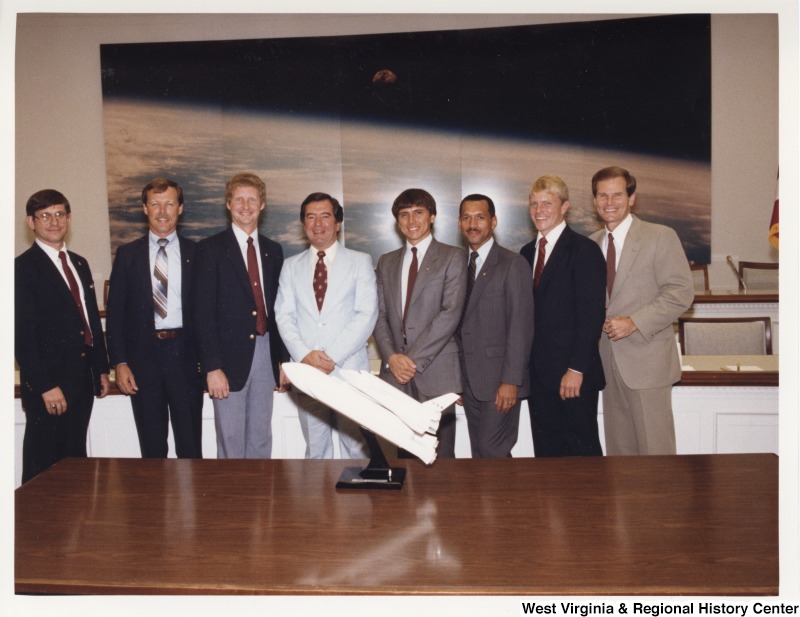 Fourth from the left, Representative Nick J. Rahall (D-W.Va.) with six unidentified members of the crew of a space shuttle. On the far right, stands Representative Bill Nelson (D-Fla.). A small space shuttle is on the table in front of the men is a small space shuttle.