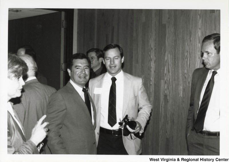 Representative Nick J. Rahall (D-W.Va.) smiles for a photograph in a group of unidentified people moving around at an event.