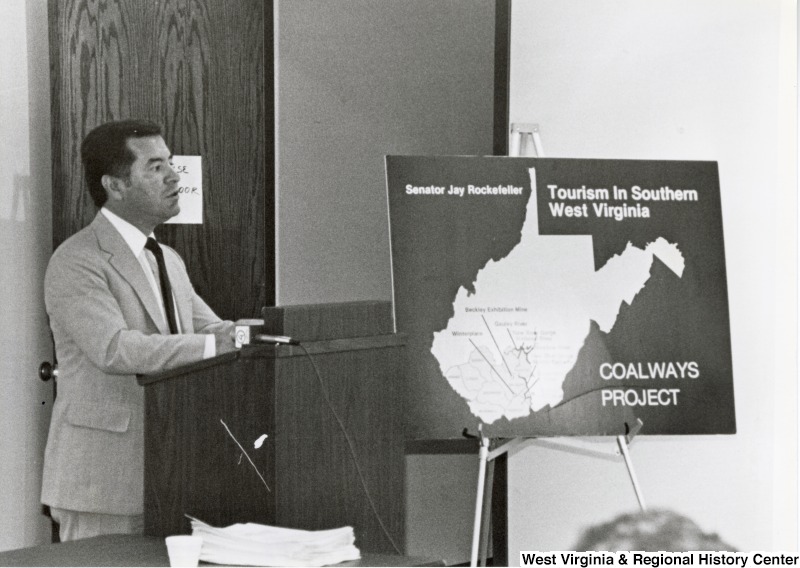 Representative Nick J. Rahall (D-W.Va.) gives a speech behind a podium. A sign showing a picture of West Virginia that reads "Senator Jay Rockefeller Tourism in Southern West Virginia Coalways Project."