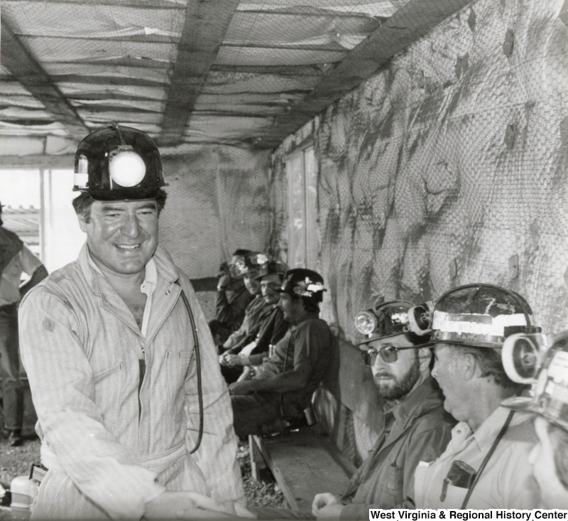 Representative Nick J. Rahall (D-W.Va.) is seen wearing a mining uniform and headlamp surrounded by unidentified coal miners.
