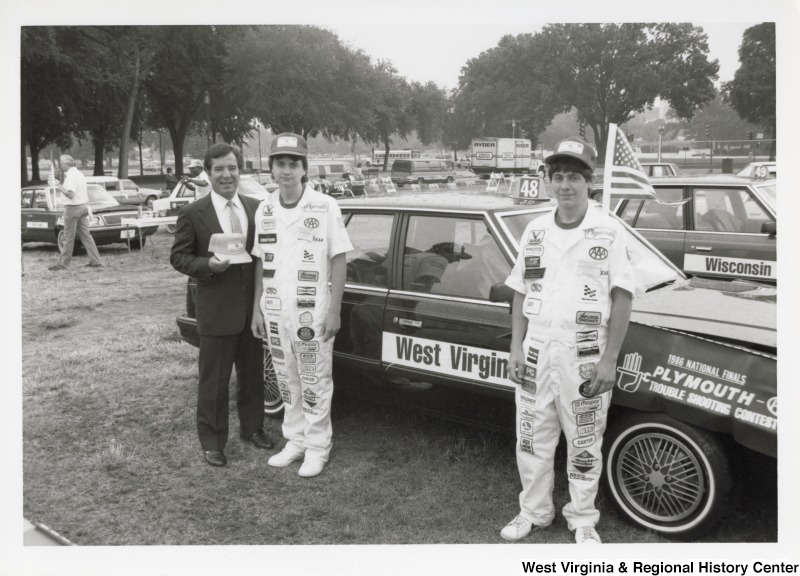 On the far left, Representative Nick J. Rahall (D-W.Va.) holds a hat and smiles for a photograph with two unidentified car drivers at the Plymouth Trouble Shooting Contest.