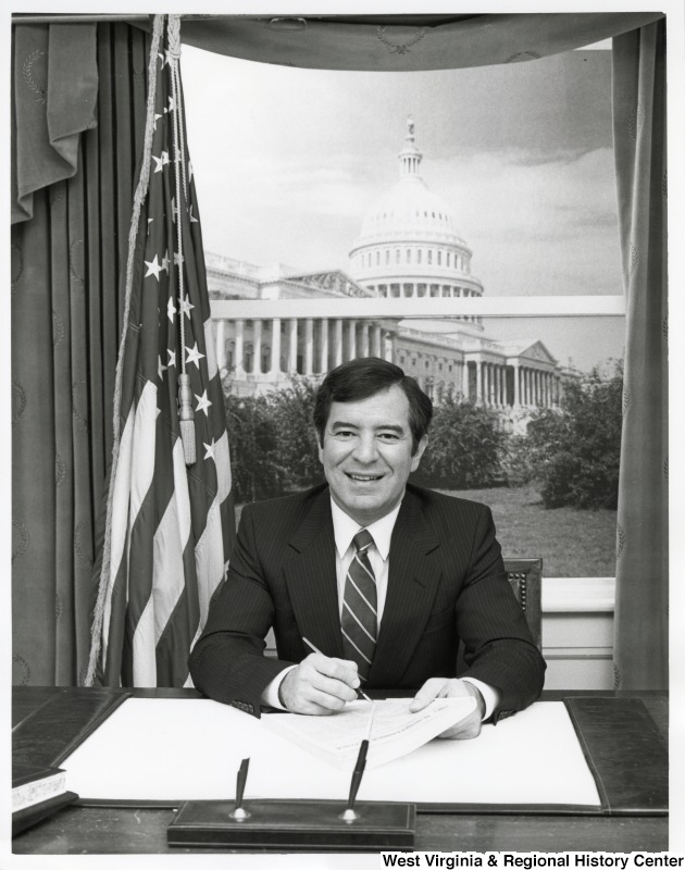 Representative Nick J. Rahall (D-W.Va.) is seated at a desk signing papers, with the Capitol building visible in the background.