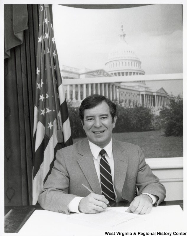 Representative Nick J. Rahall (D-W.Va.) seated behind a desk, holding papers to sign with the Capitol building in the background.