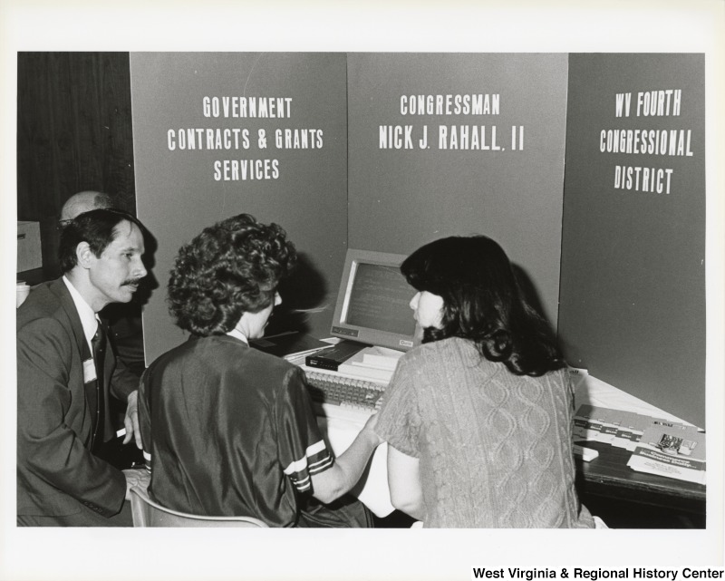 An unidentified man and two unidentified women sit around a computer with a sign that reads, "Government Contracts and Grants Services Congressman Nick J. Rahall, II WV Fourth Congressional District."