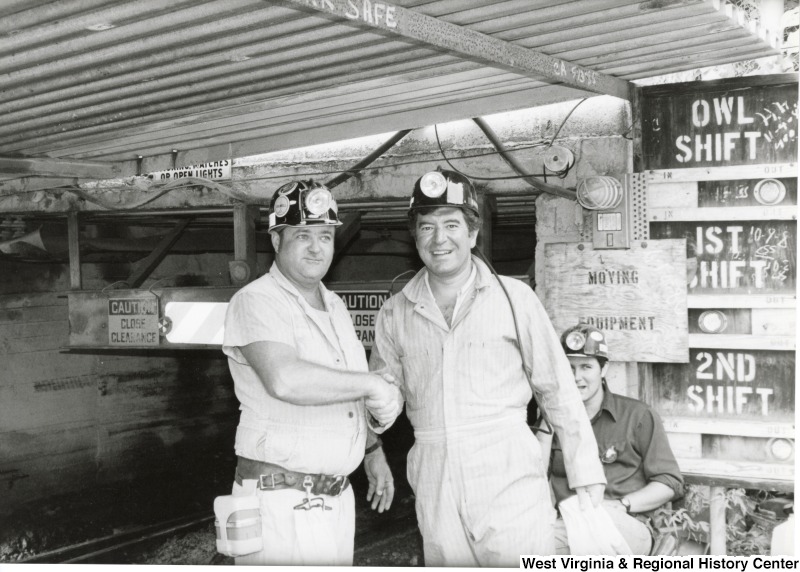 Congressman Nick Rahall dressed in a coal miner uniform, shaking hands with an unidentified coal miner at Old Ben Coal Mine. A second unidentified coal miner is watching in the background.