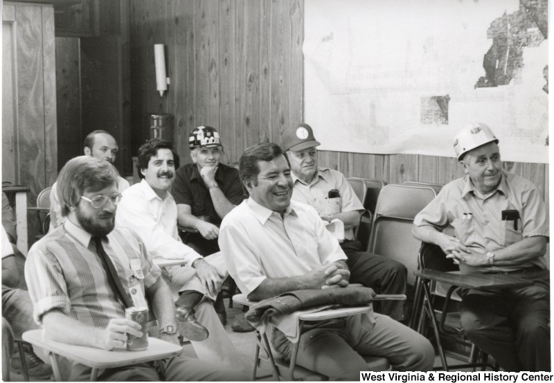 Congressman Nick Rahall sitting in a meeting room with Old Ben Coal Mine miners and Bob Stone, Secretary-Treasurer of the United Mine Workers of America.  Bob Stone is seated in the back with the wearing a hard hat with stickers on it.