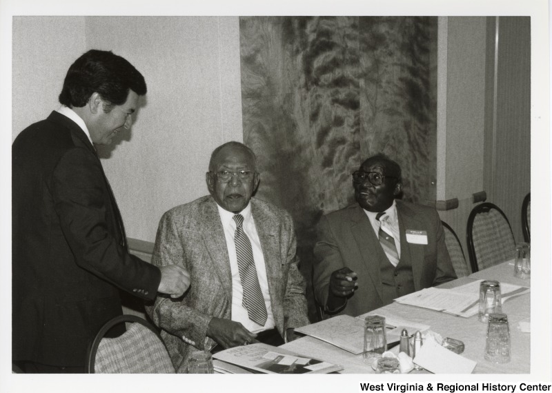 Congressman Nick Rahall speaking with two unidentified men sitting at a table during an Economic Development Seminar in Bluefield, West Virginia.