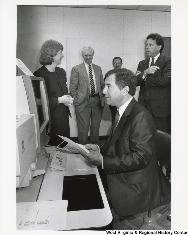 Congressman Nick Rahall looking at a computer during a visit to Marshall University. There is an unidentified group of people in the background.