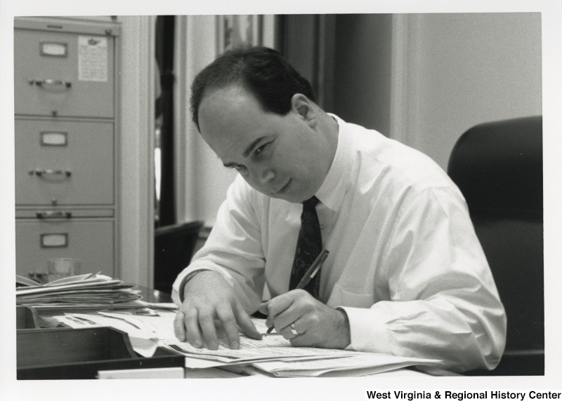 An unidentified man sits behind a desk writing on papers.
