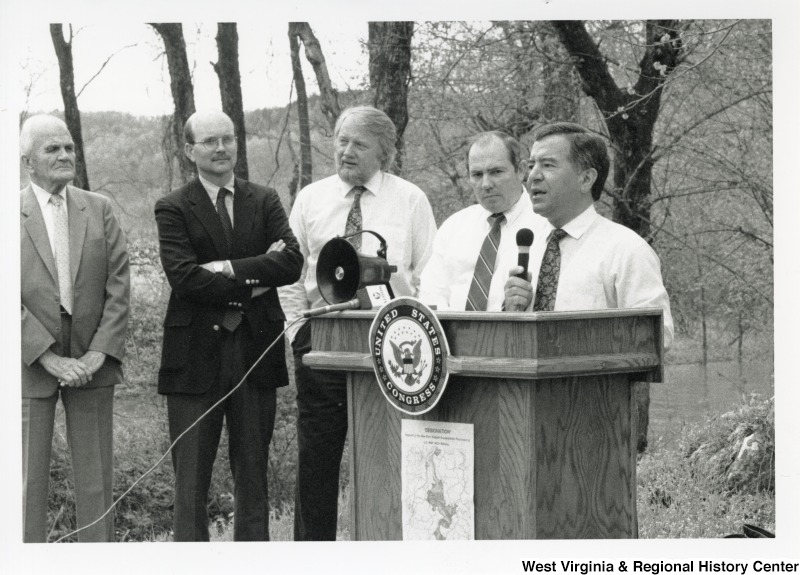 On the right, Representative Nick J. Rahall (D-W.Va.) gives a speech behind a podium in the presence of four unidentified men.