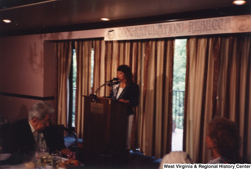 An unidentified woman stands behind a podium and gives a speech under a banner that reads "Congratulations! Rebecca" at Rebecca Rahall's graduation party.