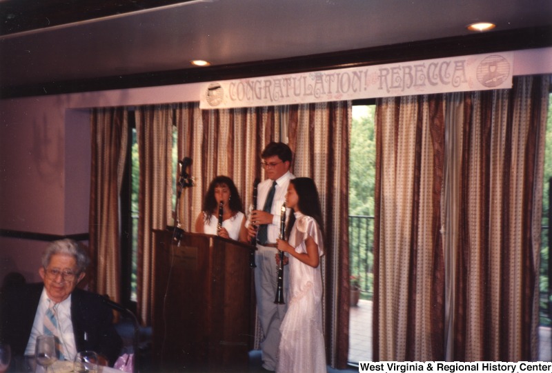 Two unidentified young women and an unidentified young man stand around a podium under a banner that reads "Congratulations! Rebecca" and play clarinets.