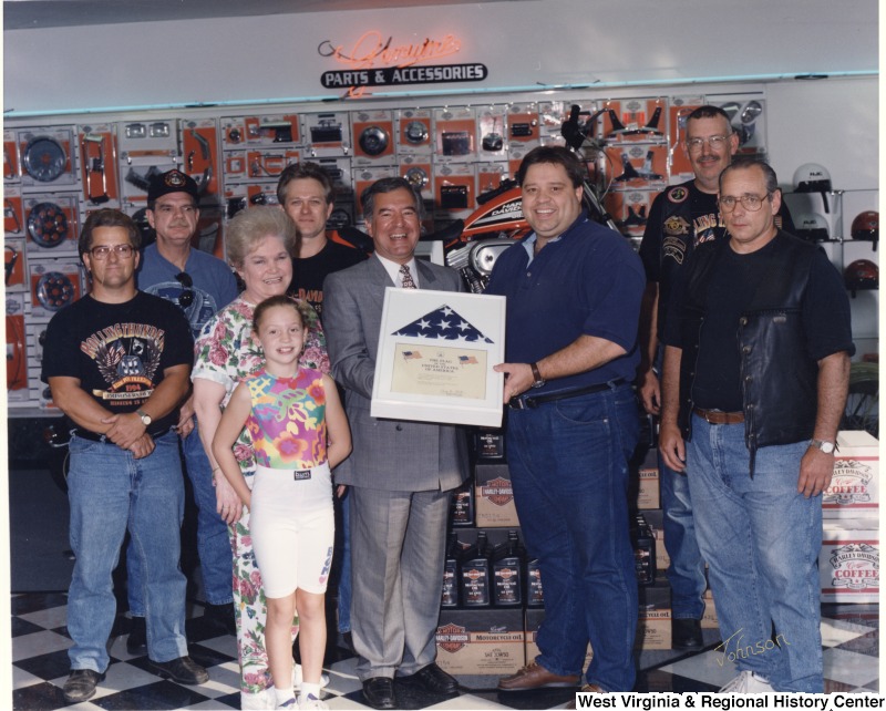 In the middle, Representative Nick J. Rahall (D-W.Va.) stands and holds a framed American flag for a photograph with a group of eight unidentified people at Benjy's Harley Davidson shop.