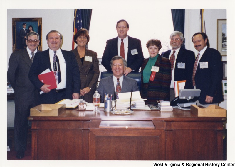 Seated behind his desk, Representative Nick J. Rahall (D-W.Va.) smiles for a photograph with the West Virginia Housing Authority. Those present in the photograph include: David Riggs, Bill Dotson, Marvin Gray, Tony Buzzi, Rossmeyer, Judith Priest, and Manuel Cartelle.