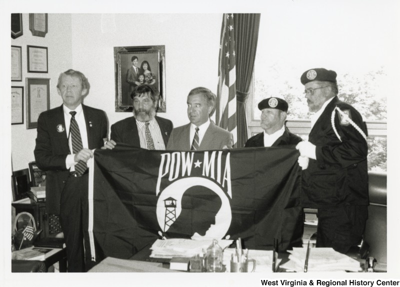 In the center, Representative Nick J. Rahall (D-W.Va.) stands between four unidentified men, two in military uniforms, and holds a POW-MIA flag in his office.