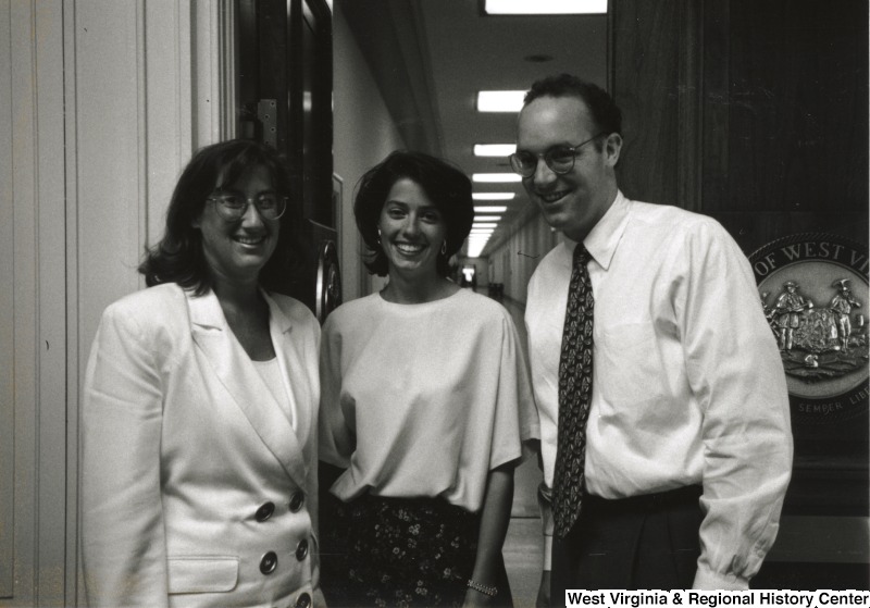 Two unidentified women and an unidentified man stand in a hallway with the Seal of West Virginia off to the side.