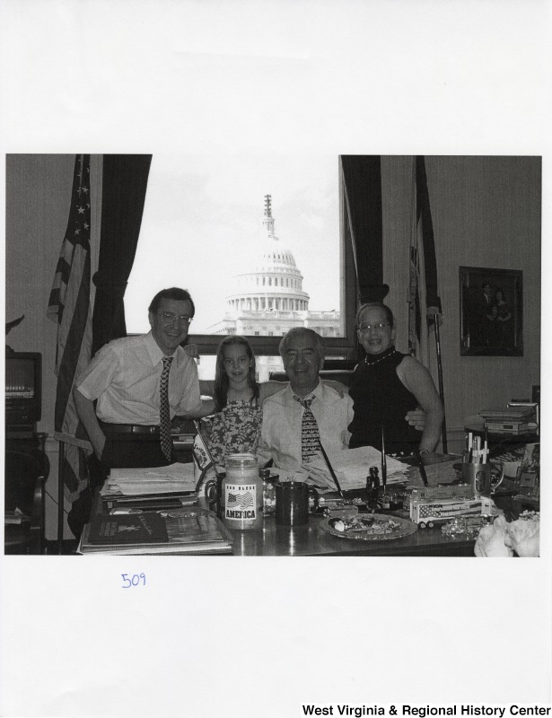 Second from the right, Representative Nick J. Rahall (D-W.Va.) sits behind his desk and smiles for a photograph with two unidentified young girls and an unidentified man.