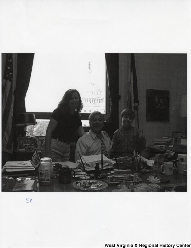 Representative Nick J. Rahall (D-W.Va.) sits at his desk for a photograph between an unidentified woman and an unidentified boy.