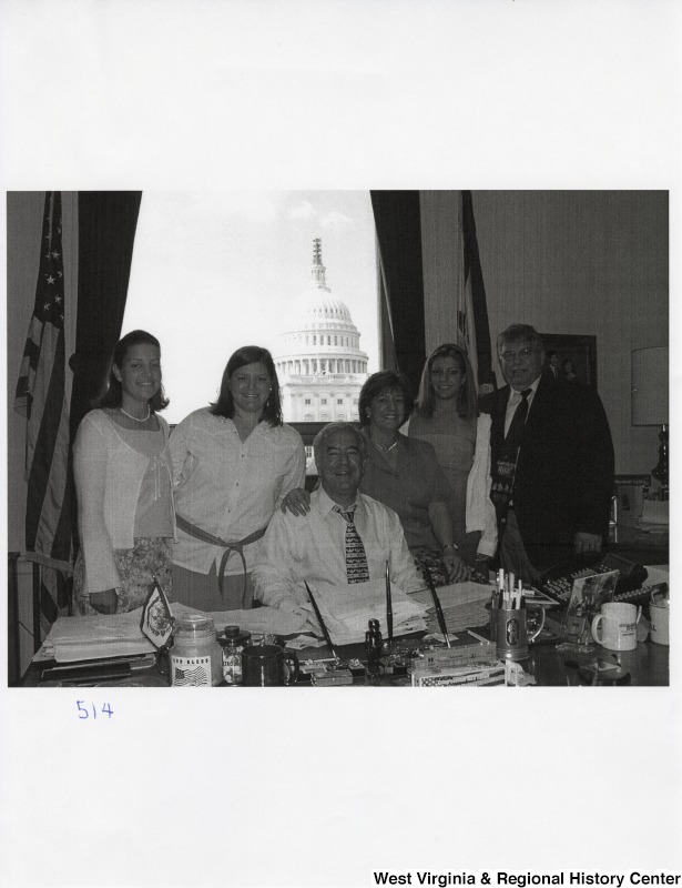 Representative Nick J. Rahall (D-W.Va.) sits at his desk for a photograph between a group of four unidentified women and an unidentified man.