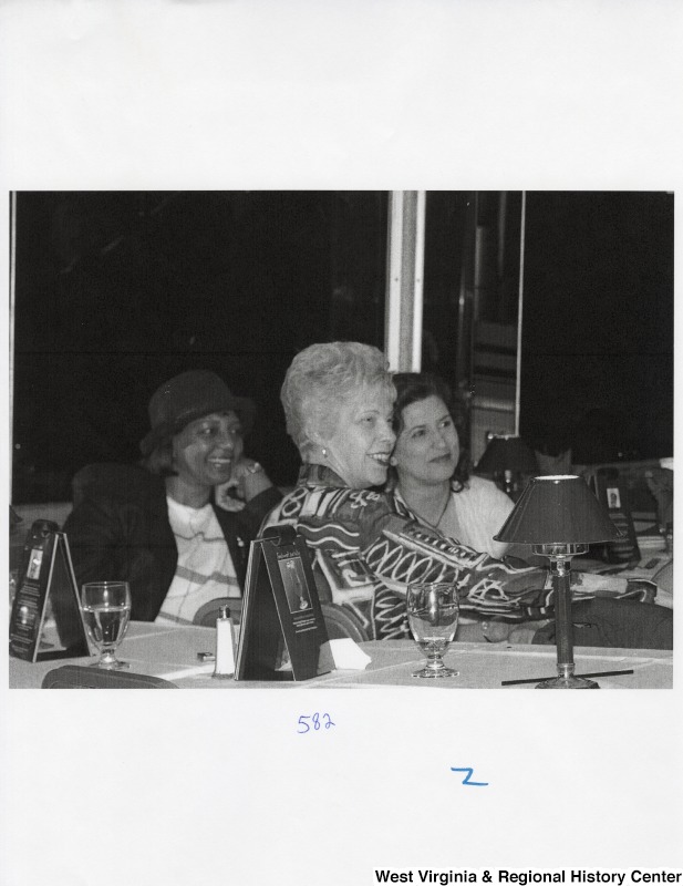 Three unidentified women watch something off to the right from a group of tables at a party.