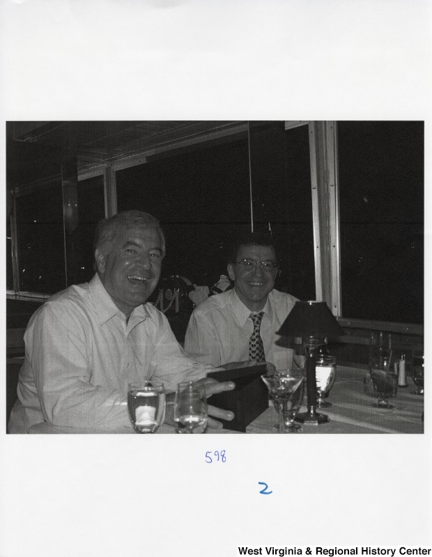 On the right, Representative Nick J. Rahall (D-W.Va.) sits with an unidentified man at a table during a party.
